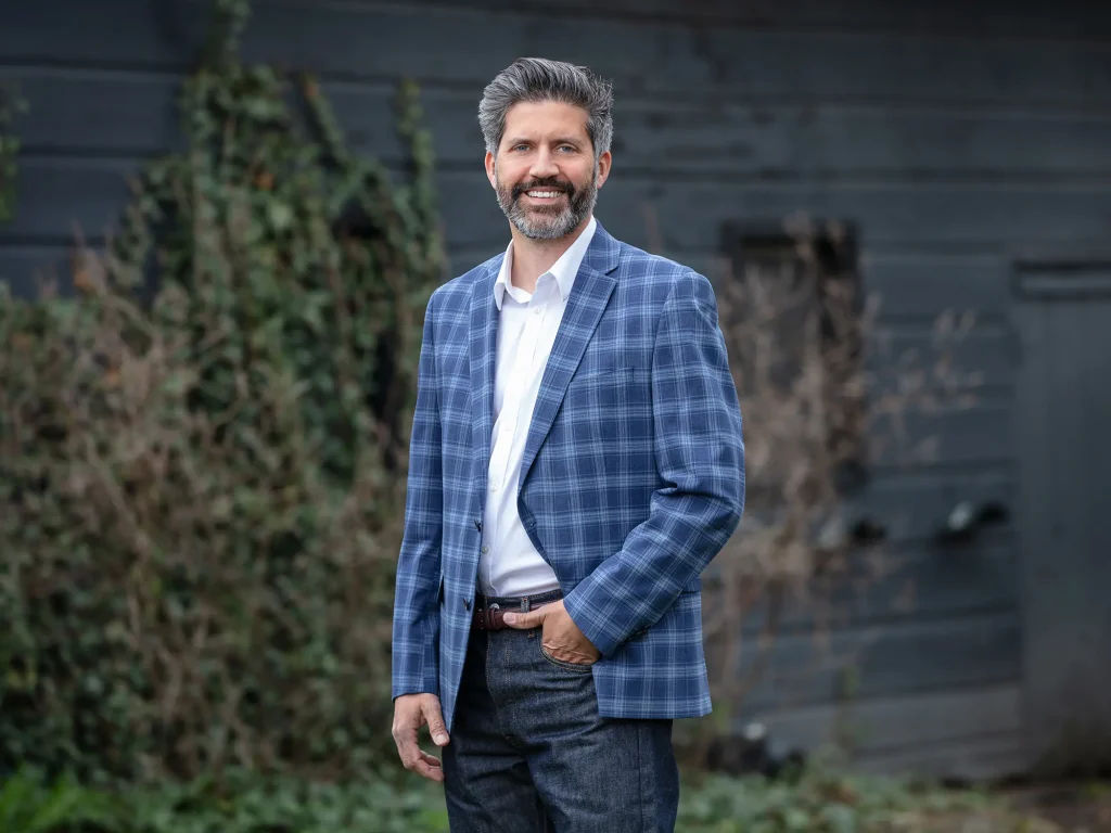 Smiling man in a blue plaid blazer stands outdoors against a dark wooden wall.